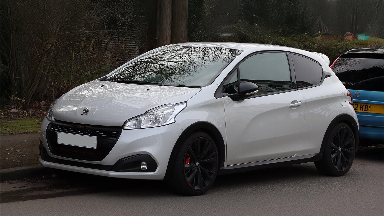 Peugeot 208 blanche, 3 portes, jantes noires, design sportif, stationnée en bord de route.