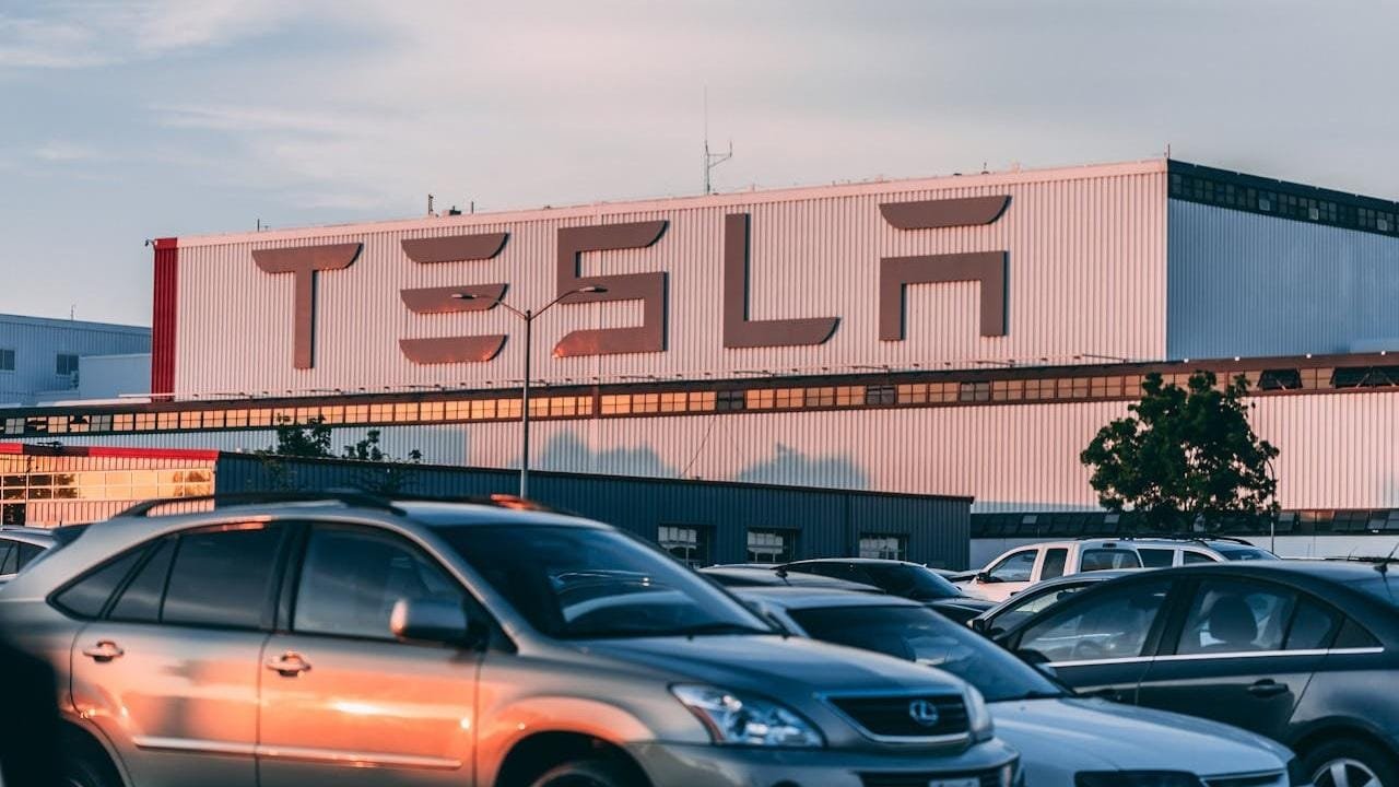 Usine Tesla avec son logo en grand sur le bâtiment, entourée de voitures sous le soleil couchant.