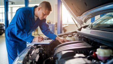 Un mécanicien en combinaison bleue examine le moteur d'une voiture tout en prenant des notes sur son bloc-notes.